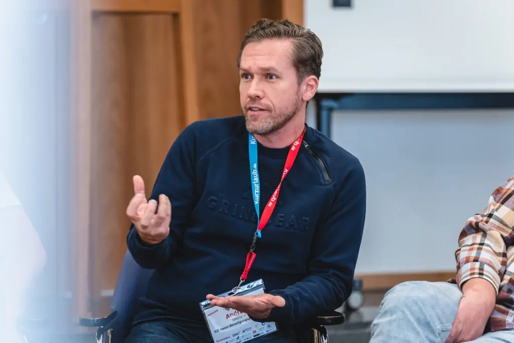 Dittlmann-Partner-ICC Ein Mann mit kurzen Haaren und Bart, der ein blaues Sweatshirt und ein rotes Schlüsselband trägt, gestikuliert beim Sprechen. Er sitzt in einem Raum mit Holzpaneelen, die an einen Konferenzbereich in einem gehobenen Hotel erinnern, und verkörpert die Haltung eines erfahrenen Beraters, der sein Publikum anleitet.